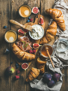 Breakfast with freshly baked croissants, ricotta cheese, figs, fresh berries, prosciutto, honey in glass jar and coffee espresso over rustic wooden background, top view, vertical composition