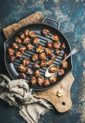 Flat-lay of roasted chestnuts in cast iron grilling pan over rustic wooden board and dark blue...
