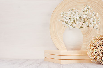 Soft home decor of beige bamboo dish, twigs and white small flowers in ceramic vase  on white wood background. Interior.