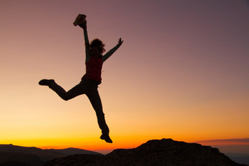 Silhouette of happy joyful young attractive woman jumping and having fun at the mountain against the sunset. Freedom, adventure and leisure vacation concept.
