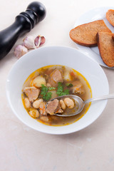 Bean soup in white plate with metal spoon, several toast on white plate on a light stone background.