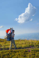Father with baby in backpack carrier is hiking in mountains. Mountain landscape with Totoro cloud shape on blue sky.