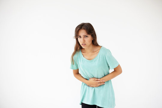Picture of casually dressed young brunette woman holding hands on her belly while suffering from stomach pain or having menstruation cramp, looking at camera with painful expression on her face