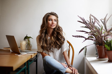 Portrait of thoughtful attractive young female freelancer with loose wavy hair using generic laptop for distant work, sitting at cafe table by window and looking outside, having pensive look