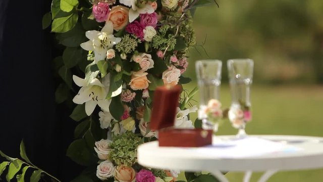 Wedding rings of white gold in the wooden box on the table. Glasses decorated with rorses and berries. Decorations for outdoor ceremony.
