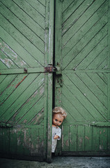 The old doll peeks out from behind the doors. Old green wooden shed. Fear, hate, horror, loneliness.