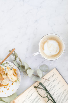 Superficie de m&aacute;rmol con capuccino, un libro, unas gafas de lectura y reposter&iacute;a