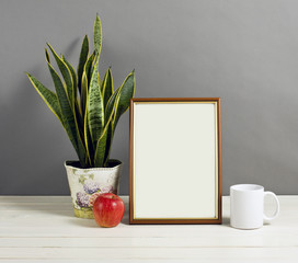 Mock up of blank photo frame with plant pot, mug and apple on wooden shelf.