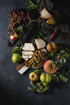 Serving board with sliced camembert cheese and baked bunch of green grapes served with bread, glass of red wine, corkscrew, apples, pears, leaves over black texture background. Top view with space