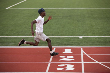African male sportsman in a cap fast running short distance on stadium track, he trains speed