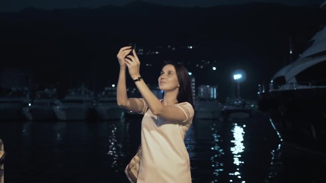 Yong Woman Taking Pictures By Smartphone. Stylish Summer Traveler Woman With Phone Outdoors In European City, Night Bay With Yachts On The Background