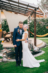 Stylish happy smile bride with a bouquet of peonies and groom hold in hands and go. Arranged five fingers. On the background on the nature in the courtyard of the house. 