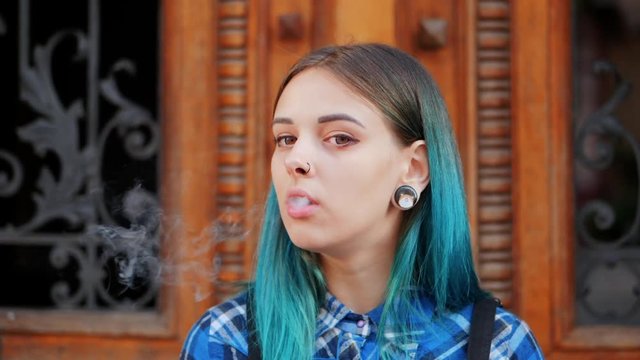 Young punk girl smoking cigarette by the wooden door.Hipster with blue dyed hair. Woman with piercing in nose, violet lenses, ears tunnels and unusual hairstyle.Slow motion.