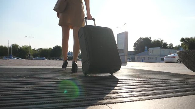 Business lady going to taxi parking from the airport with her luggage. Girl in heels stepping and roll suitcase on wheels. Woman walking with her suitcase along street. Travel concept. Slow motion