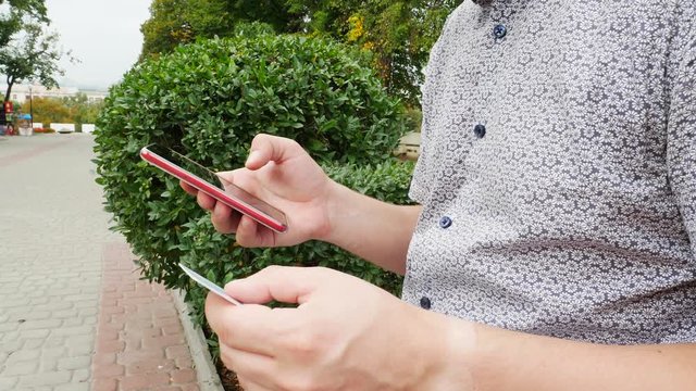Man hands holding red smartphone and credit or debit card and buy online outside in park on background of green bushes