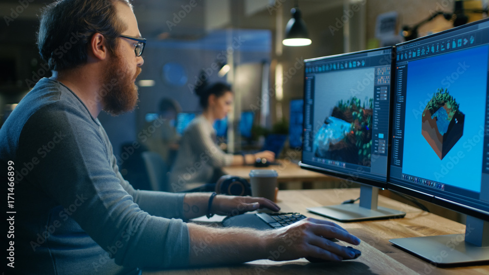 Poster Creative Male Game Developer Works with Graphics for the New Level Design, He Sits at His Wooden Table Working on Two Display Personal Computer. Other People Work in This Creative Studio.