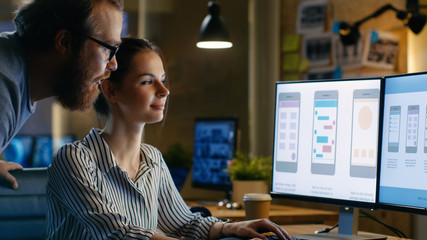 Female UX Architect Has Discussion with Male Design Engineer, They Work on Mobile Application Late at Night, She Drinks Coffee. In the Background Wall with Project Sticky Notes.