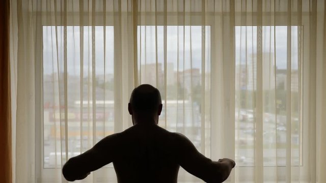 Adult man does morning exercises stretches hands in front of window. Indoors locked shot.