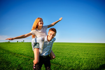 Fototapeta premium Smiling Man is holding on his back happy woman, who pulls out her arms and simulates a flight against the background of the blue sky and the green field