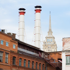 Fototapeta na wymiar Mix of two pipes and buildings of different times against sky. Old Industrial building of factory from red brick, smoking chimneys, tower residential building, ecology