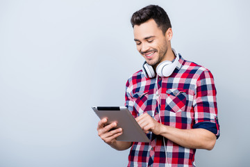 Successful young brunet freelancer hipster in checkered shirt is standing on the pure grey background with tablet, smiling, looking at it