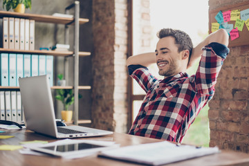 Cheerful young brunet guy is watching at his laptop screen, at his work place, with arms behind the...