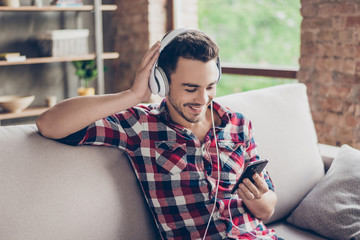 Close up portrait of attractive serene hipster dj in ckeckered shirt, enjoys the stereo sound in big modern ear phones, touches them, smiling. He is at home, loft designed room, free time, rest