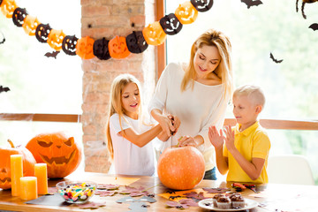 Cheerful mom with two small adorable kids preparing the jackolantern at decorated room at home loft style, desk top with yellow candles, sweets, fall leaves, little bats on windows