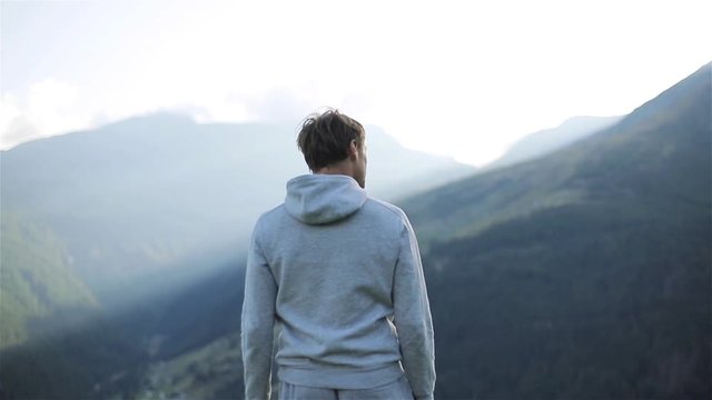 Man stands back on mountain top enjoying view pointing showing finger slow motion. Smiling happy male silhouette in grey sportswear looking at valley landscape and at camera. Winner training travel