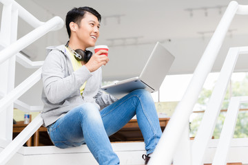 Happy young Asian employee drinking coffee while using a laptop in the interior of a modern office