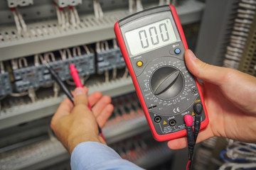 Multimeter in hands of electrician close-up on blurred background of electrical elements. engineer performs adjustment work in control cabinet of industrial equipment