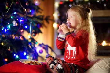 Happy little girl having milk and cookies by a fireplace in a cozy dark living room on Christmas eve