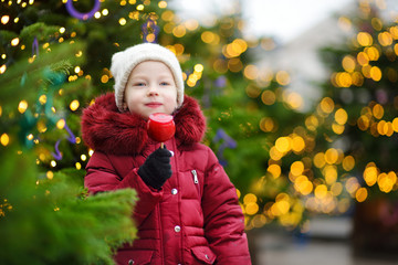 Adorable little girl eating red apple covered with sugar icing on traditional Christmas market.