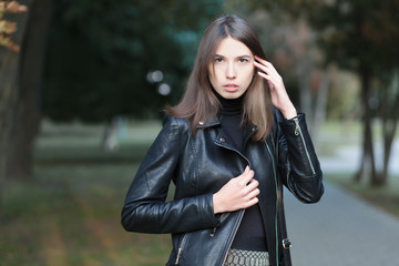 Art portrait of a young pretty brunette woman posing outdoors in black leather coat againt city park blurry background