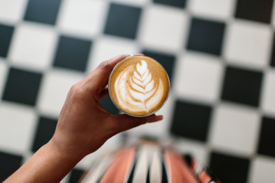 A woman holding a cortado and looking down at a checkered floor.
