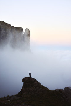 From a precarious and airy rock peak a hiker stands in owe of the majestic misty mountains that surround him.