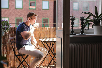 A blonde man enjoying his coffee on a sunny summer morning sitting down outside on his balcony. Photo from inside apartment looking out.