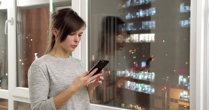 Young woman at home flat by window dialing and calling on mobile phone 4k video. Girl in apartment with night city landscape and talks on smartphone
