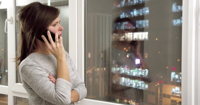 Young woman at home flat by window talks on mobile phone 4k video. Girl standing in apartment with night city landscape and calling on smartphone