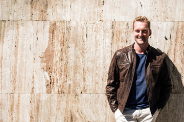 Portrait of a blonde man wearing a brown leather jacket leaning against a stone wall outdoors on a sunny summer day.