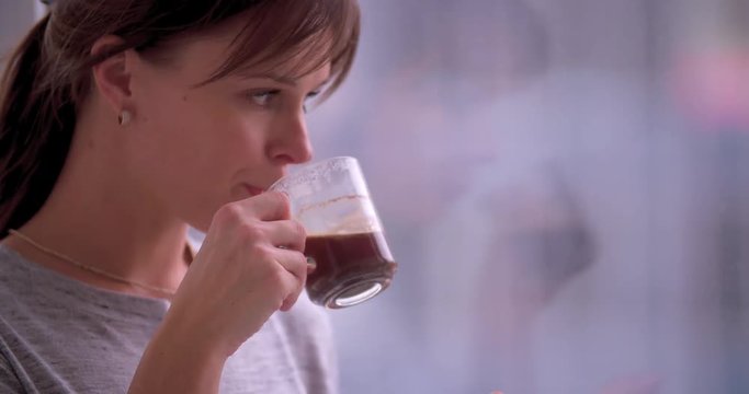 Young woman standing by window drinks coffee 4k video. Girl looking at morning or evening at home flat apartment. Close-up of hand holds cap