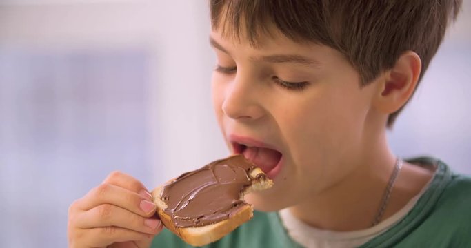 Boy child eating chocolate hazelnut spread toast HD slow-motion video. Close-up of kid hand holds cocoa sweet paste bread and bites chews. Morning breakfast food