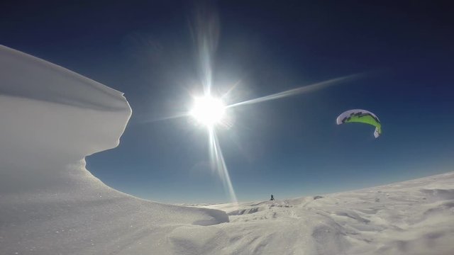 An idyllic steady shot of a man parasailing on a snowy surface. The parasail is colored white and green.