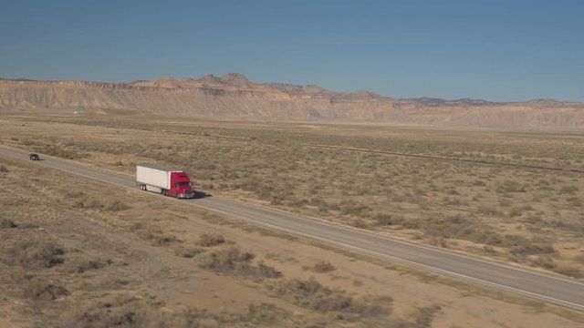 AERIAL, CLOSE UP: Flying above red freight container semi-truck driving along the interstate highway running through vast desert with sandstone rock formations in Utah, America. Lorry shipping goods