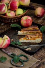 Sliced pieces of warm apple pie on rusty pan on wooden rustic table with fresh apples basket, knife, fork and sackcloth, green leaves. Autumn fruits caramelized tart with cinnamon copy space for text