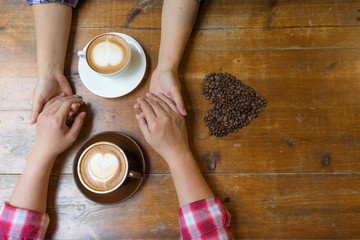 Fototapeta na wymiar Couple in love holding hands with coffee on wooden table. Photograph taken from above, side view with heart coffee
