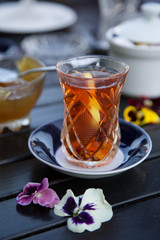 Black tea in traditional turkish and azerbaijani pear shaped glass with violet color saucer on wooden table in restaurant outdoors. Tea with sweet dessert - fruit jam in glass bowl and spoon