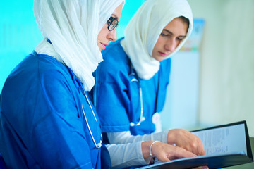 Close-up of two arabic female doctors dressed in blue uniform and white hijab sitting in a hospital reading a medical report