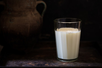 Glass of fresh farm milk with butter cookies stack on sack cloth and spoon by side, rural scene in rustic style and dark background with clay pot, organic breakfast, diary product, copy space for text