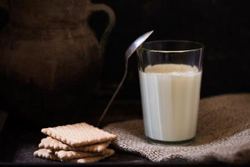 Glass of fresh farm milk with butter cookies stack on sack cloth and spoon by side, rural scene in rustic style and dark background with clay pot, organic breakfast, diary product, copy space for text